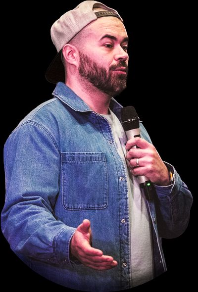 Craig Abbott talking into a microphone at a conference. He is a white male with a short black beard, wearing a denim shirt and a backwards baseball cap.